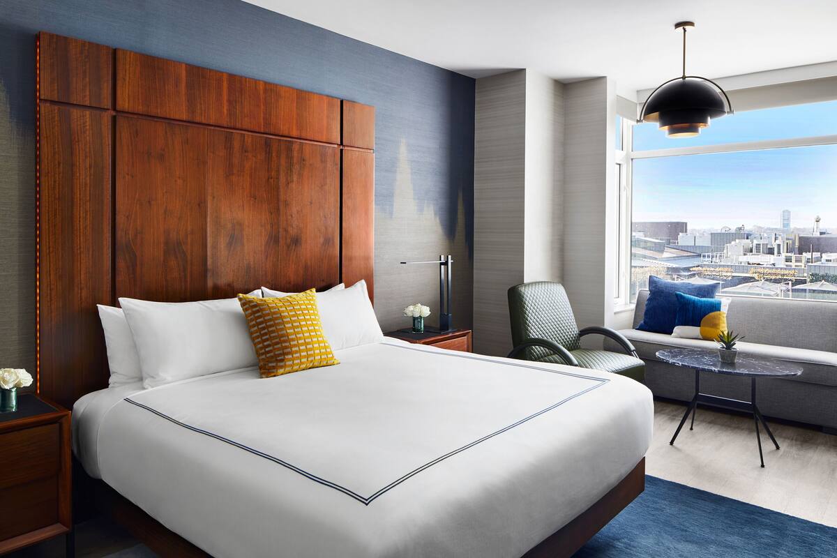 A hotel room with a large bed, a city view, a modern chair, and a small table.