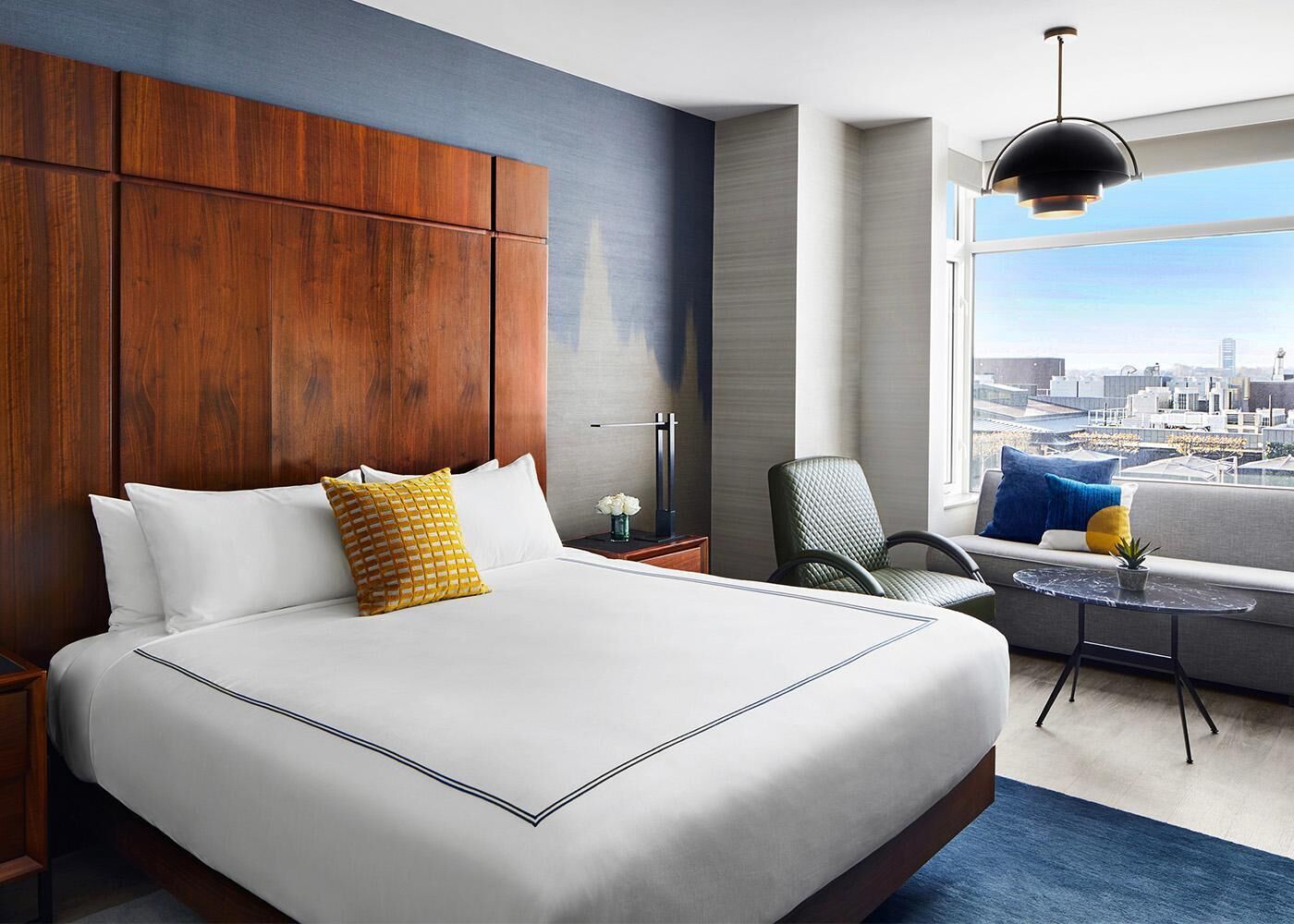A hotel room with a large bed, a view of the city, a seating area with a table, and a modern pendant light.