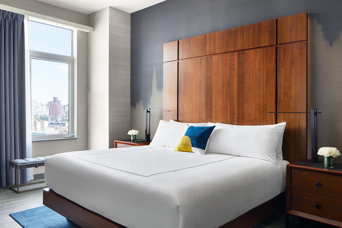 A modern hotel room with a large bed, a wooden headboard, a nightstand with a lamp, and a view of the city through the window.