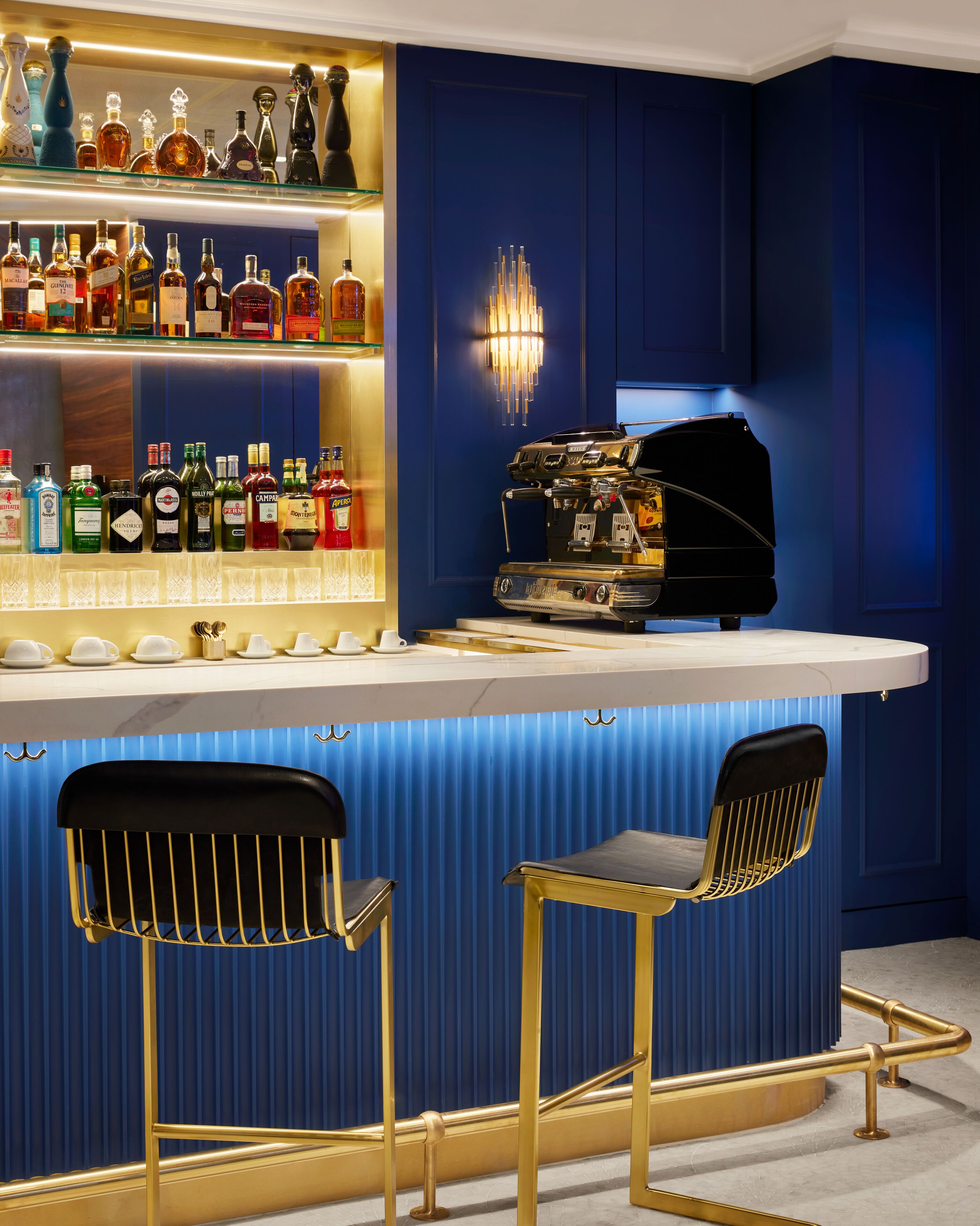 Een bar met een aanrecht, flessenkasten vol drank, twee barkrukken en een koffiemachine.
