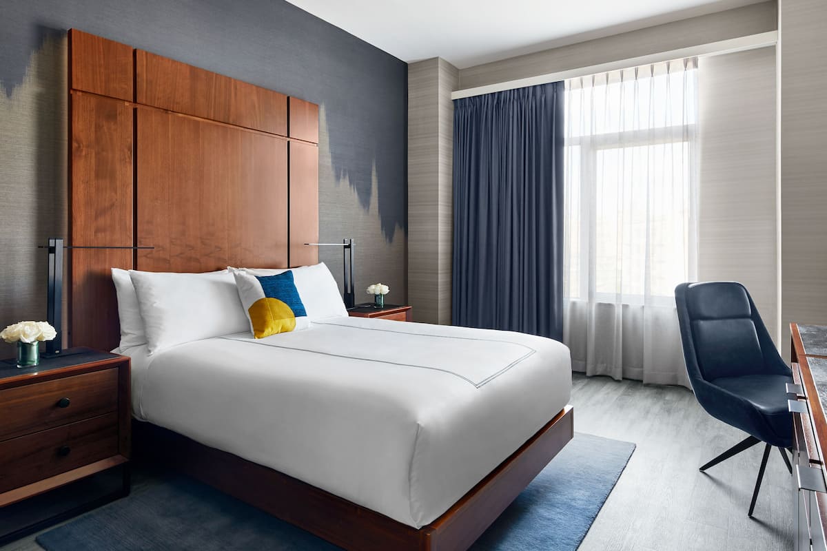 A modern hotel room with a large bed, wooden headboard, a desk with a chair, and a large window with curtains.