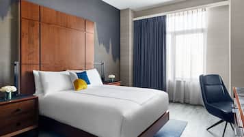 Una habitación de hotel moderna con una cama grande, cabecera de madera, un escritorio con una silla y un amplio ventanal con cortinas.