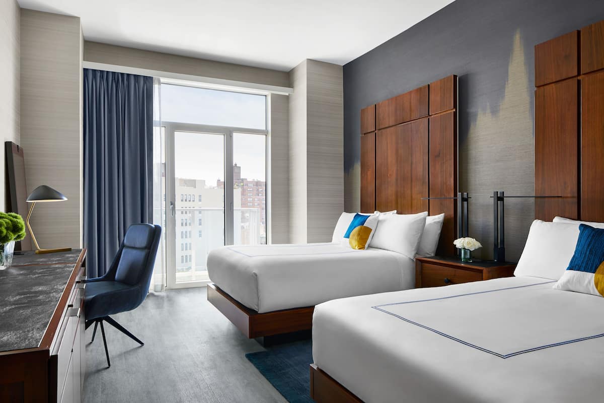 A hotel room with two beds, a desk, a chair, a large window, and a view of the city.