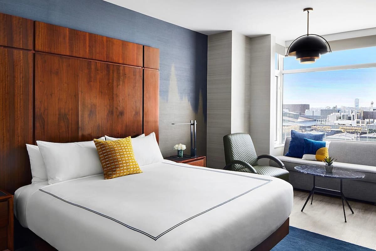 Une chambre d’hôtel dotée d’un grand lit, offrant une vue sur la ville, comprenant un coin salon avec une table et un luminaire suspendu moderne.
