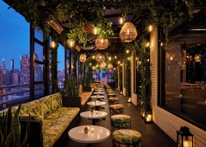 A rooftop bar with a city view, featuring a long bench seating area, round tables, and hanging wicker lamps. - Gansevoort Meatpacking (New York)
