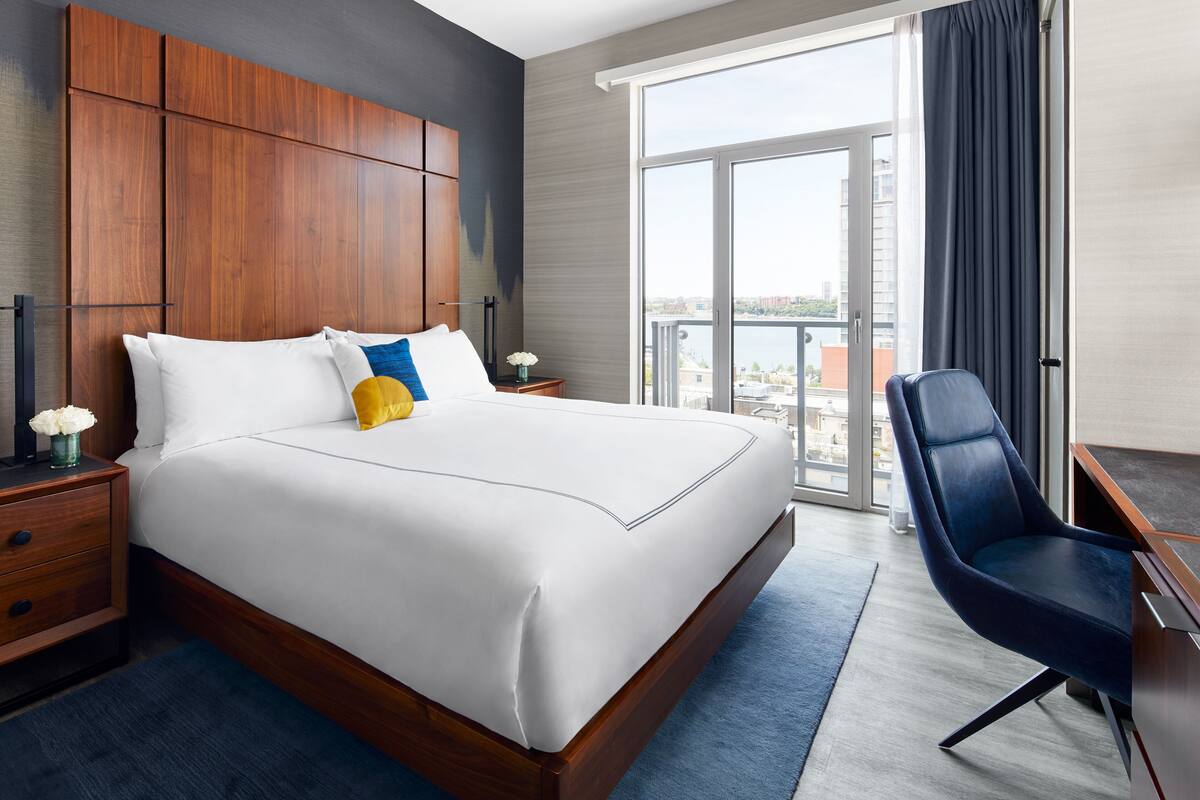 A hotel room with a large bed, a wooden headboard, a desk with a chair, and a large window with a view of buildings.