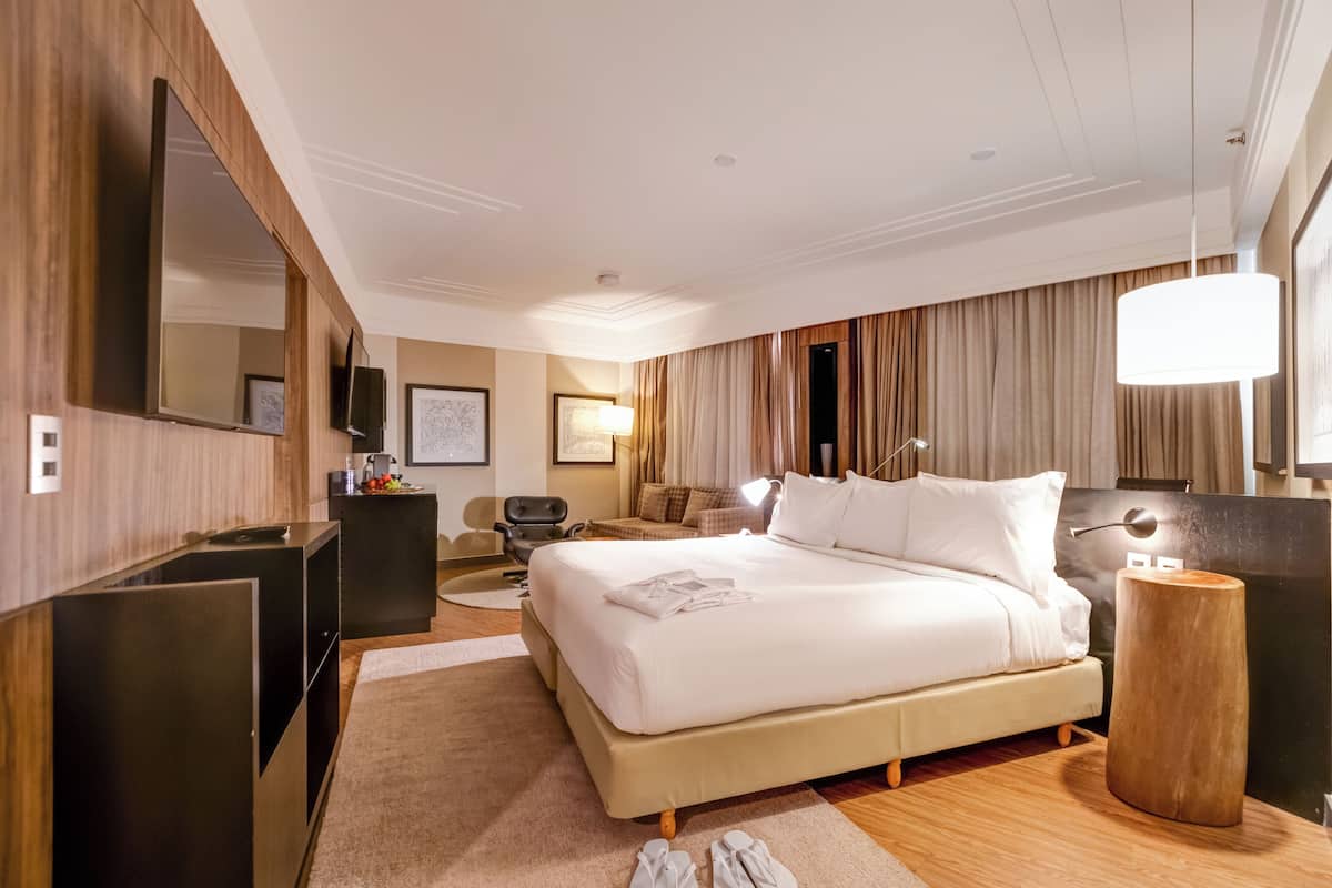 A hotel room with a large bed, a desk, a chair, a TV, and a lamp.