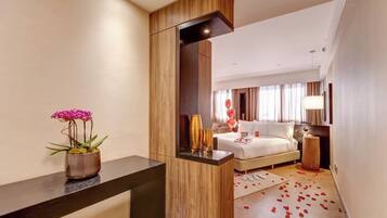 A hotel room with a bed, bedside table, mirror, potted plant, and scattered rose petals on the floor.