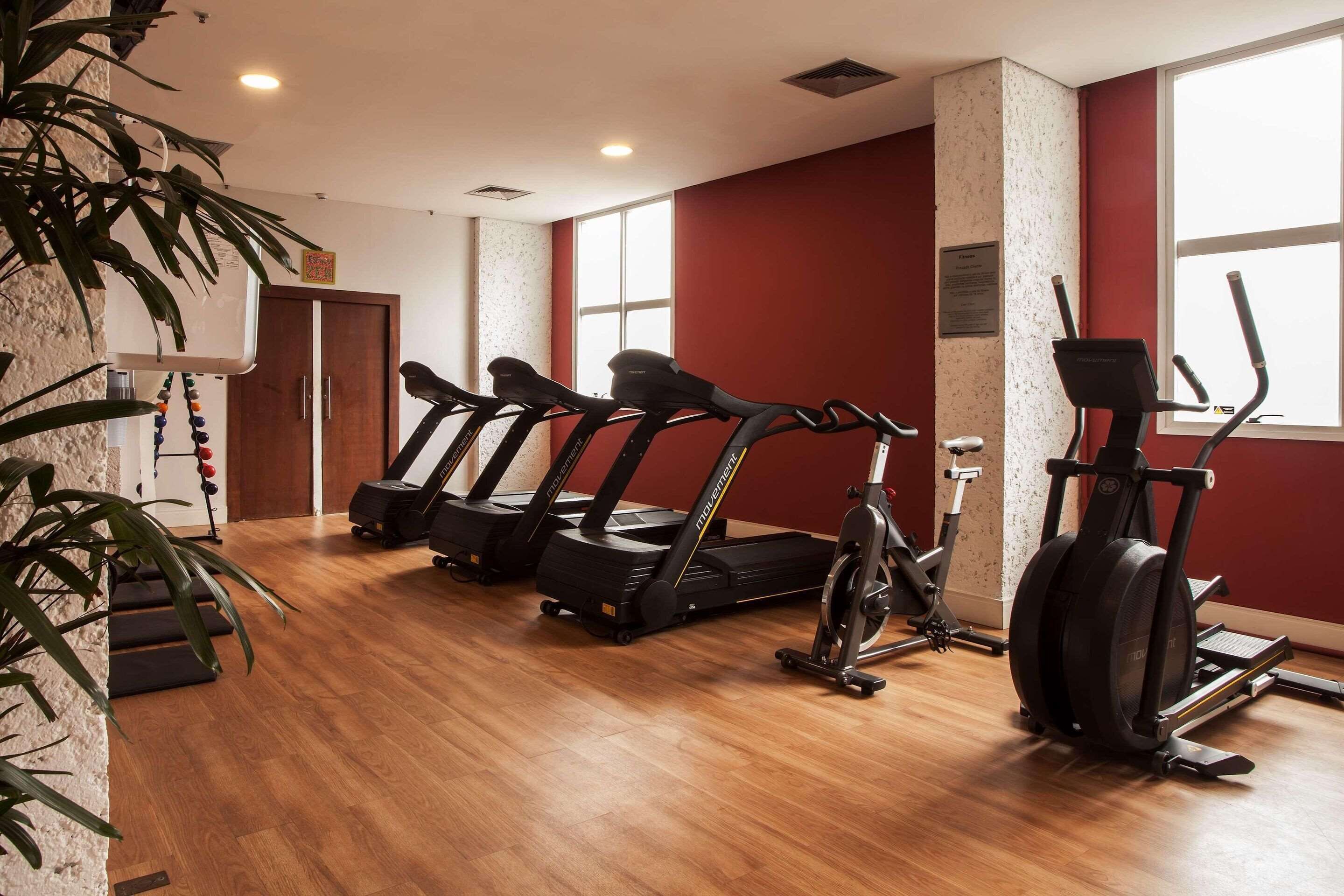 A gym with treadmills, an elliptical, and a stationary bike, wooden flooring, and a red wall.