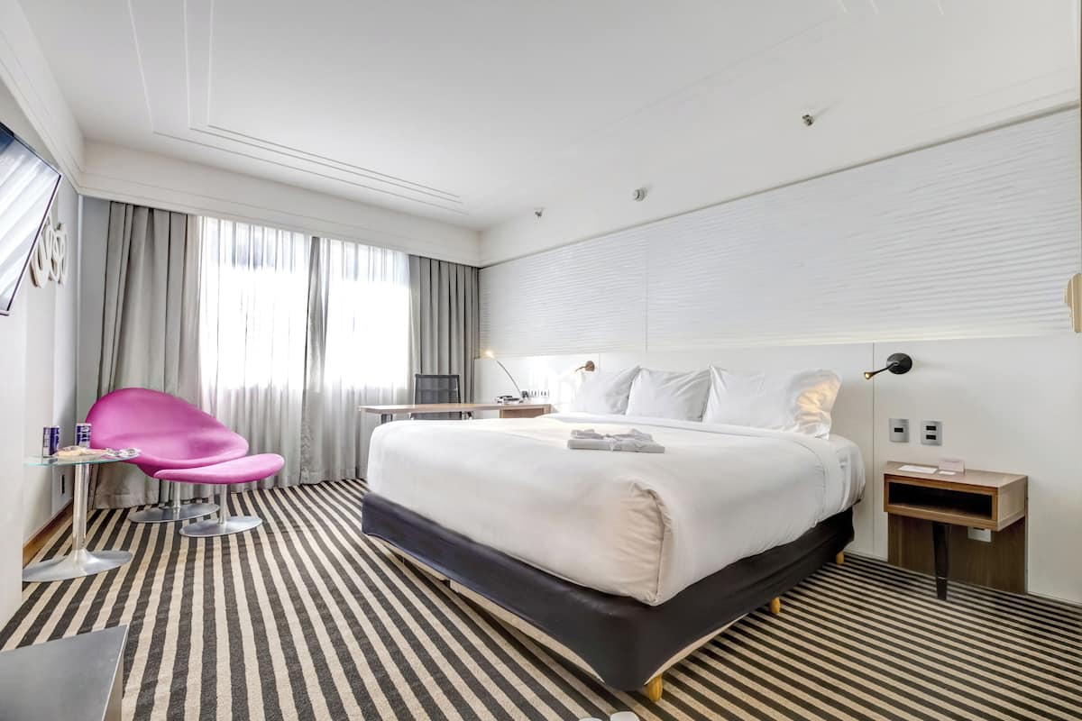 A hotel room with a large bed, a desk with a chair, a bedside table, and a window with curtains.
