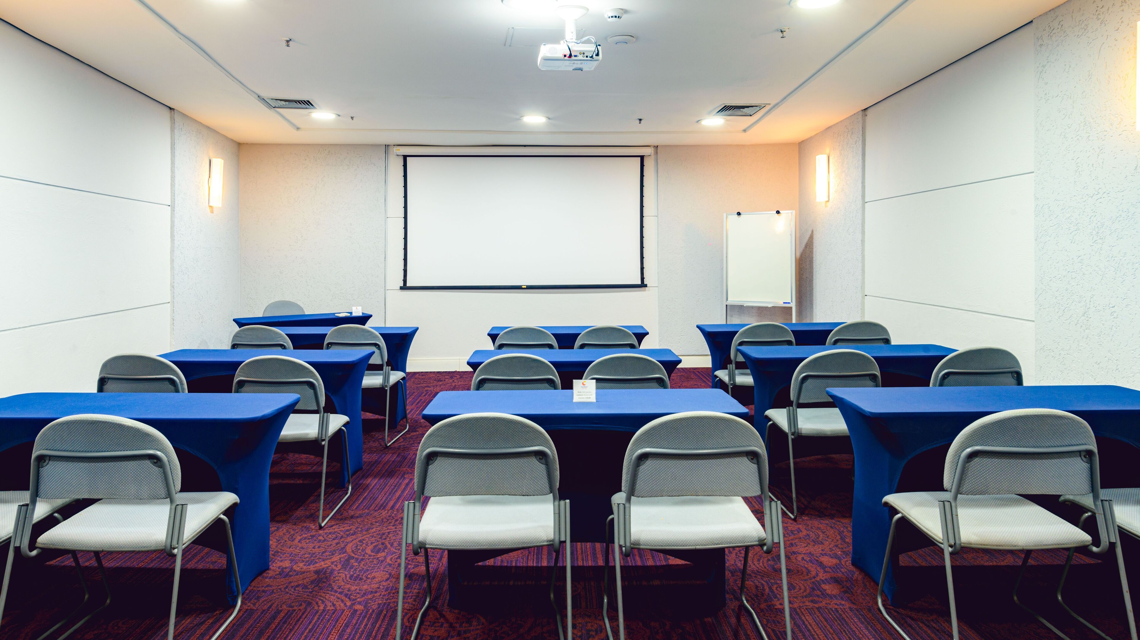 A conference room with rows of tables and chairs, a projector screen, and a whiteboard.