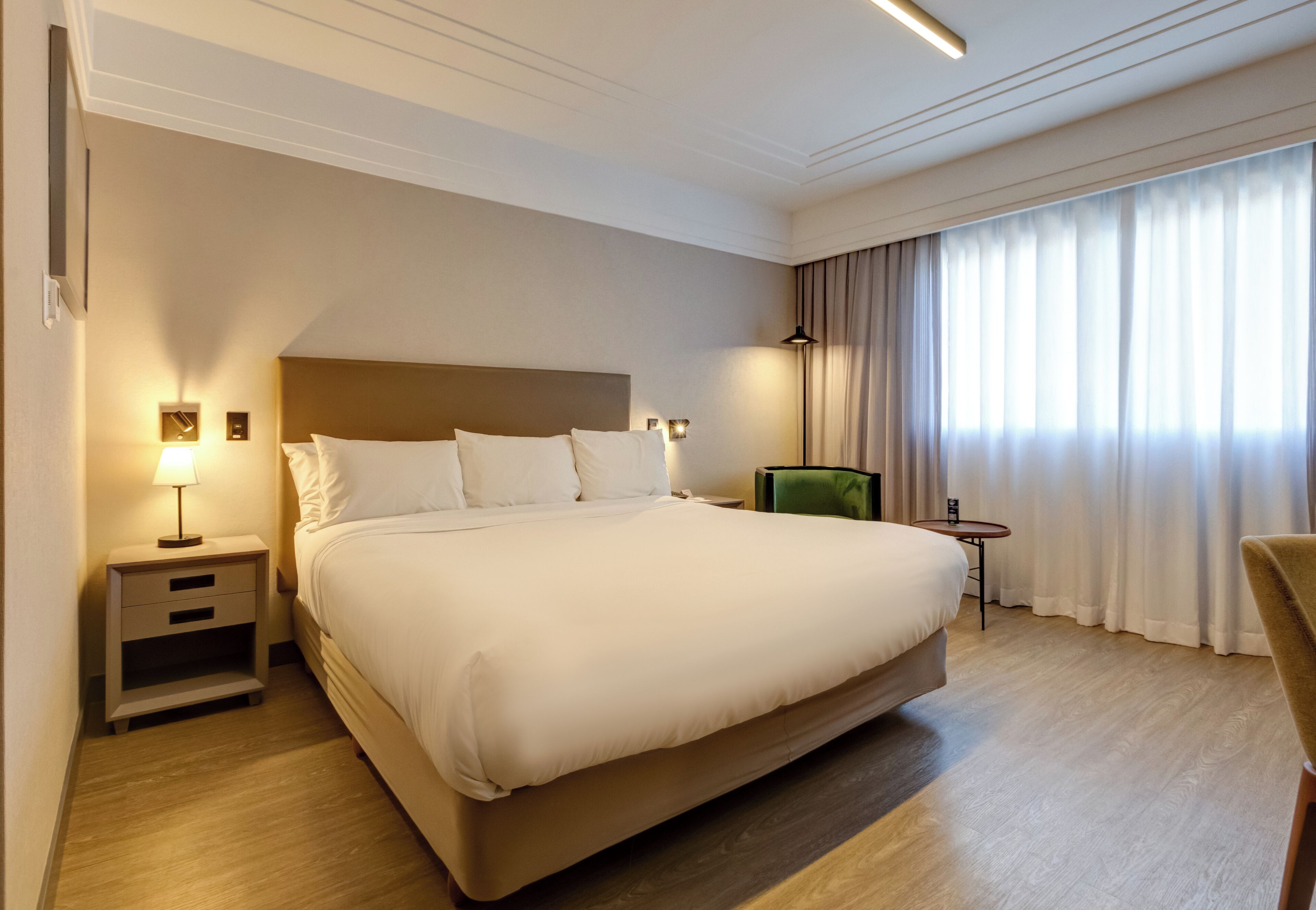 A hotel room with a large bed, bedside tables, a chair, a small table, and a window with curtains.