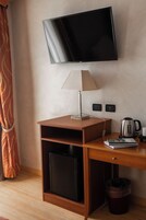 Camera d'albergo con televisione a schermo piatto fissata al muro, una scrivania in legno con una lampada, un bollitore e un cestino.
