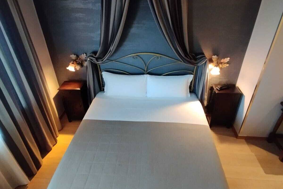 A neatly made bed with a headboard, flanked by two bedside tables with lamps, in a room with dark walls and curtains.