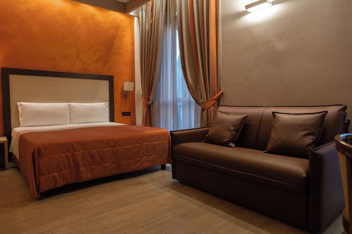 A hotel room with a bed, a sofa, and a window with curtains.
