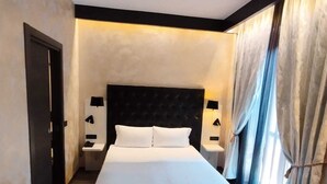 Una habitación de hotel con una cama grande, suelo de madera oscura, cabecero negro, dos mesitas de noche con lámparas y una ventana con cortinas.