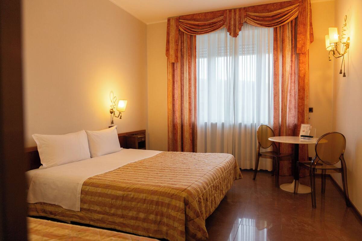 A hotel room with a bed, a desk with two chairs, a lamp, and curtains.