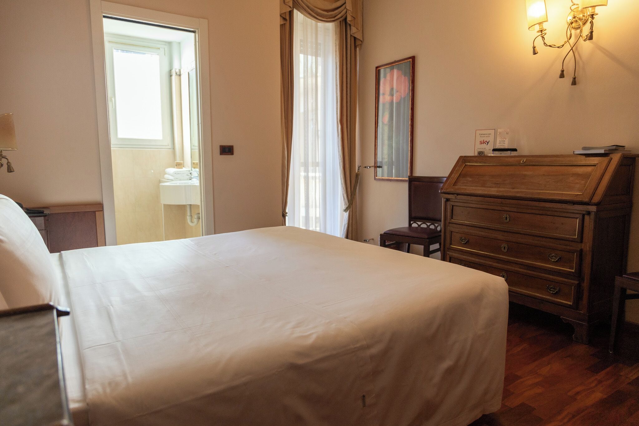 Een hotelkamer met een groot bed, een houten dressoir, een spiegel en een raam met gordijnen.