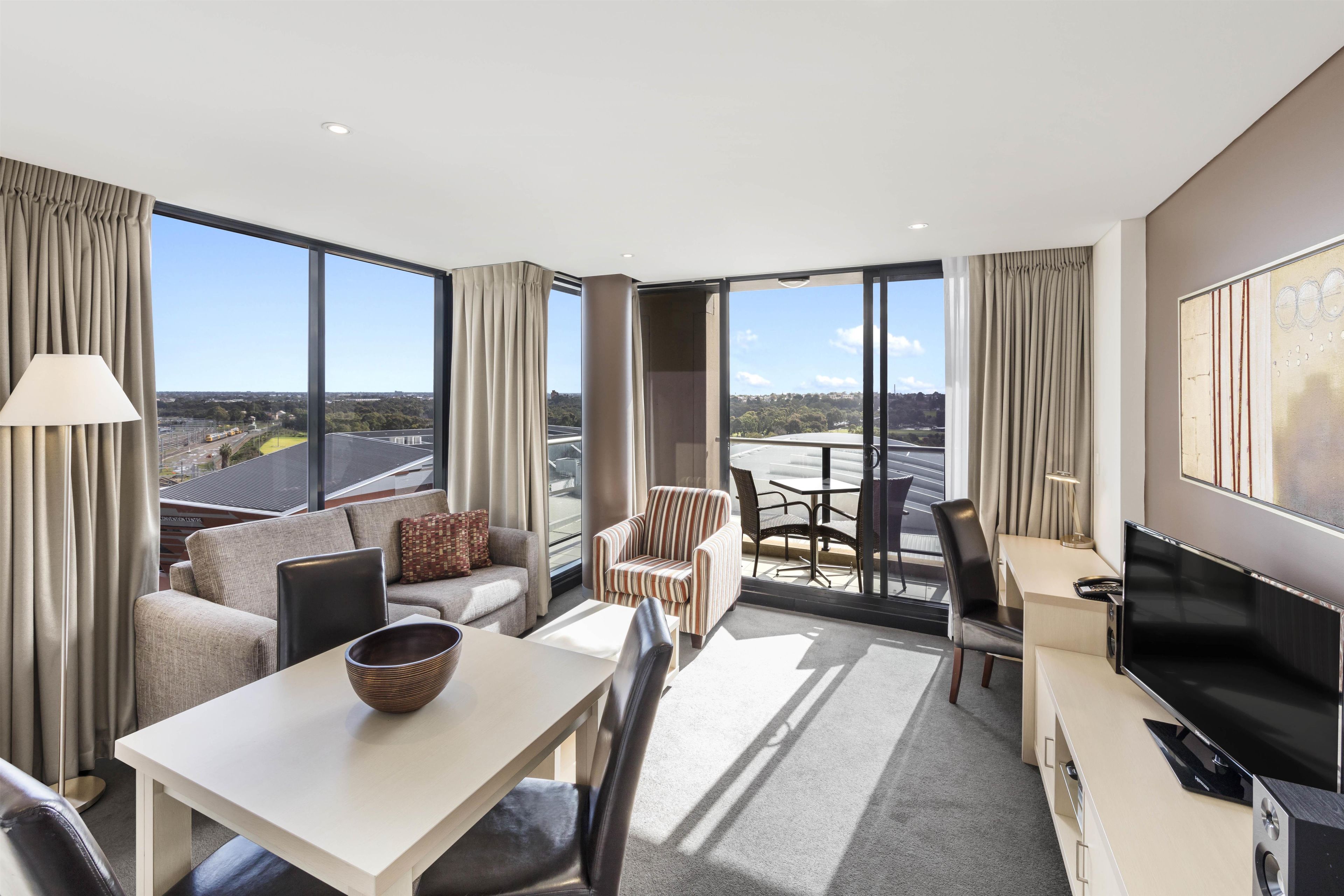 A modern hotel room with a large window, a sofa, a dining table, and a TV.