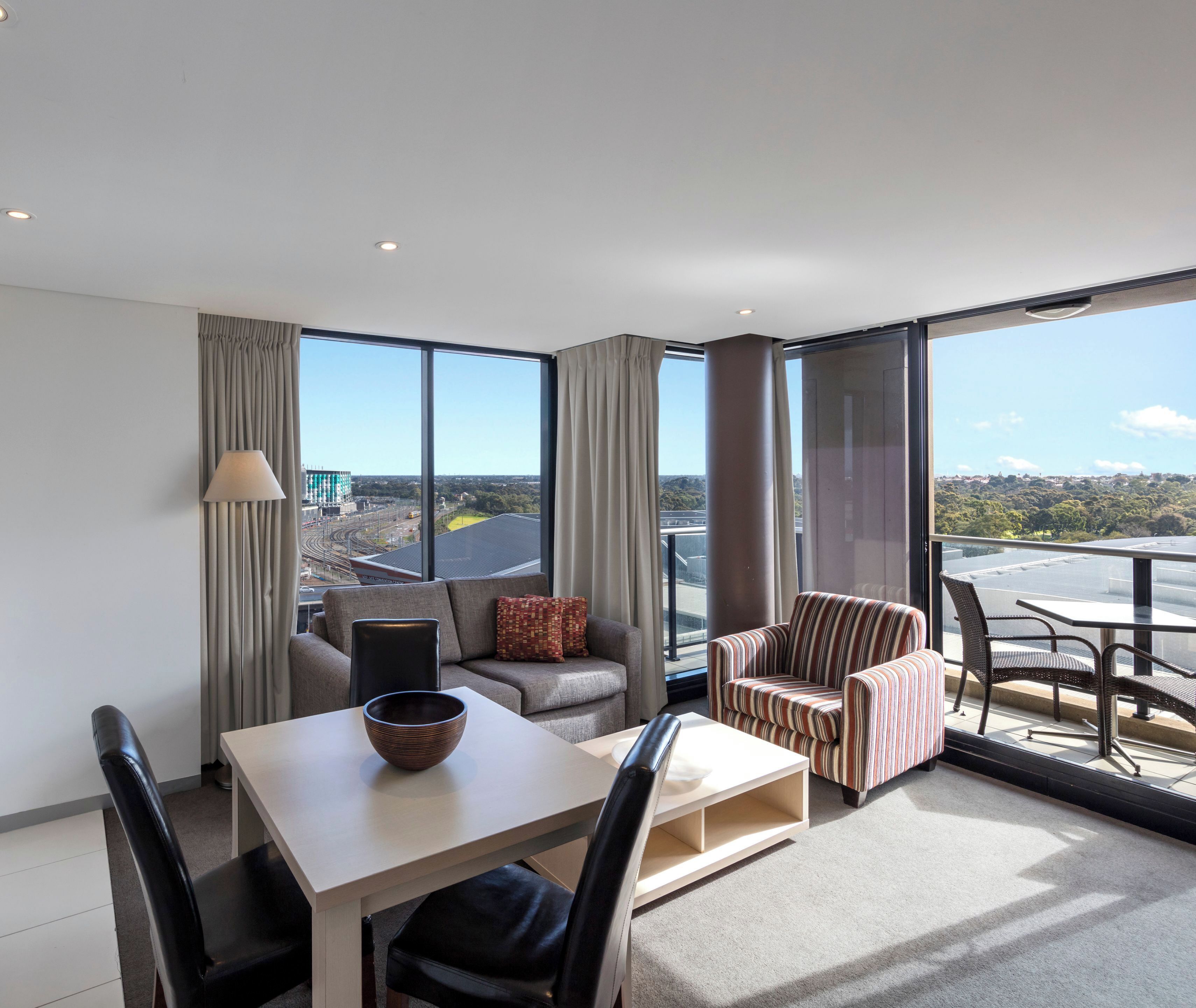 A modern hotel room with a dining area, a sofa, a striped armchair, and large windows offering a city view.