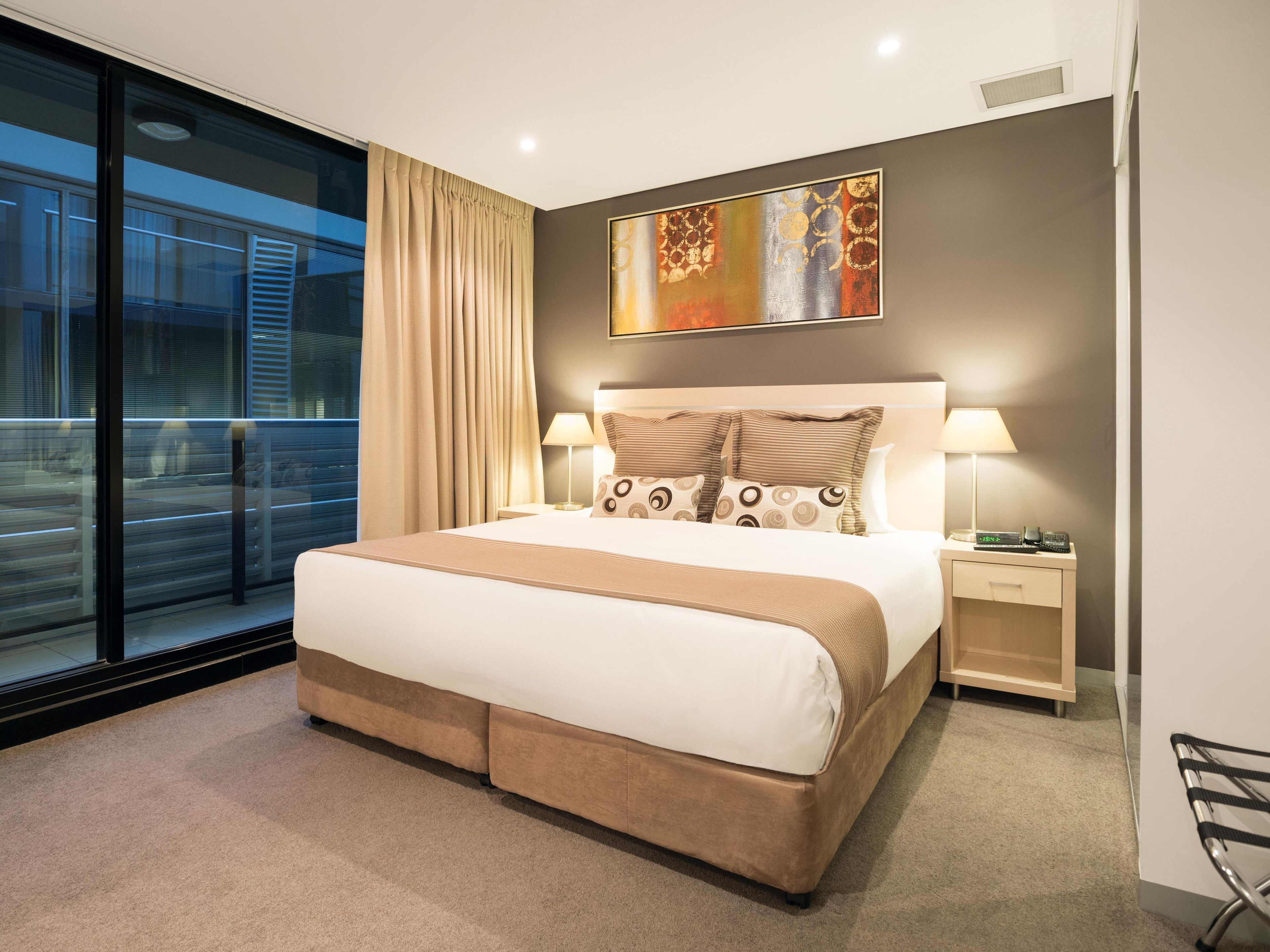 A modern hotel room with a large bed, a nightstand, a lamp, and a painting on the wall.