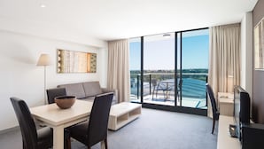 A modern hotel room with a dining area, a sofa, a TV, and a large window offering a city view.