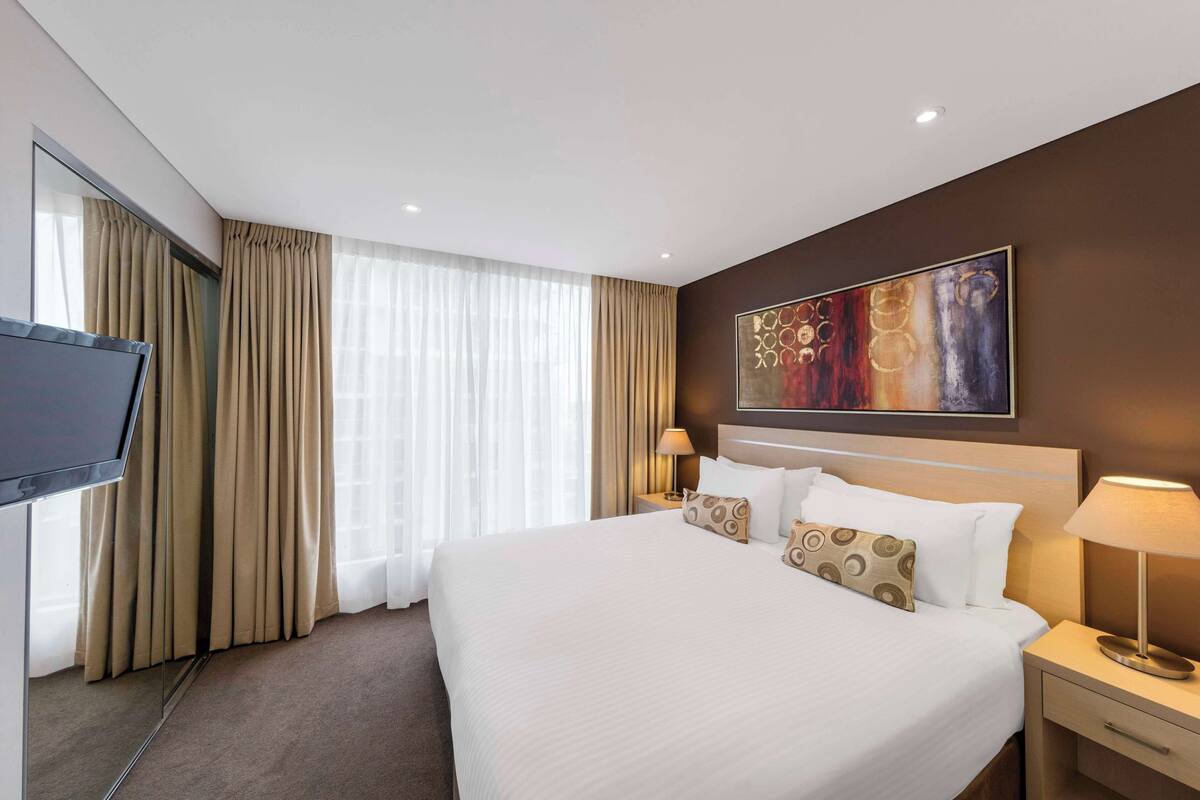 A hotel room with a large bed, a TV, a painting on the wall, and a window with curtains.