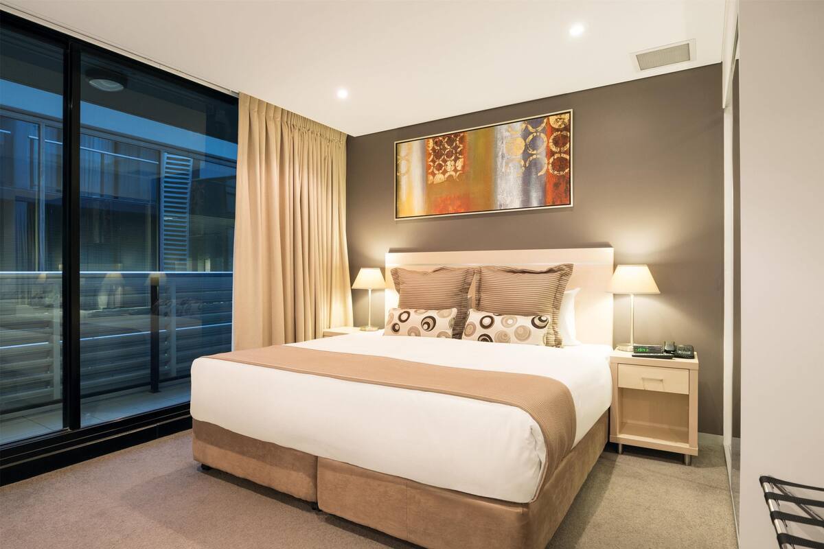 A modern hotel room with a large bed, two bedside tables with lamps, a painting on the wall, and a view of a train outside the window.