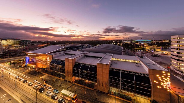 A modern building with a large glass facade and a curved roof, surrounded by a parking area and a busy street.