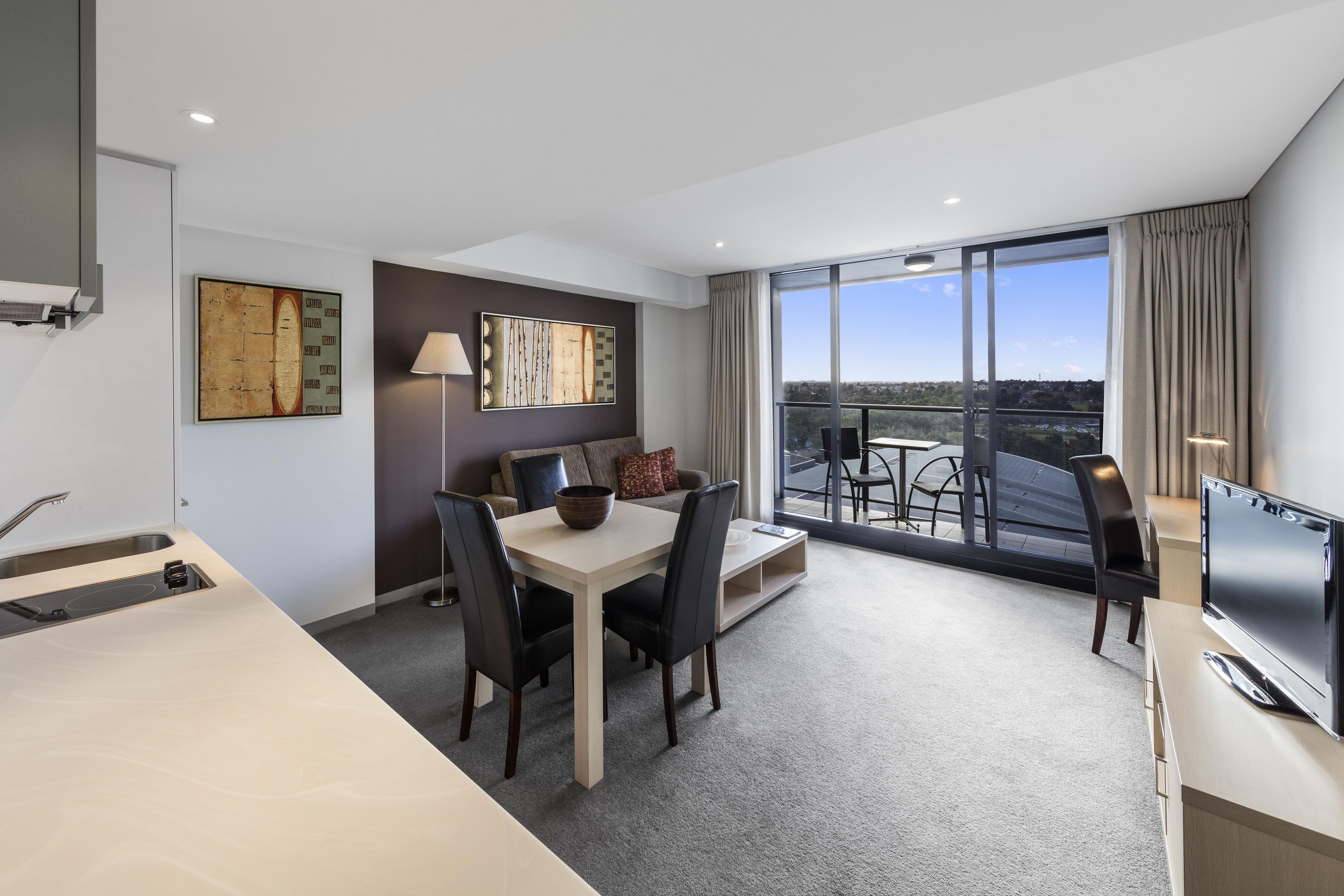 A modern hotel room with a dining area, a flat-screen TV, and a balcony with a city view.