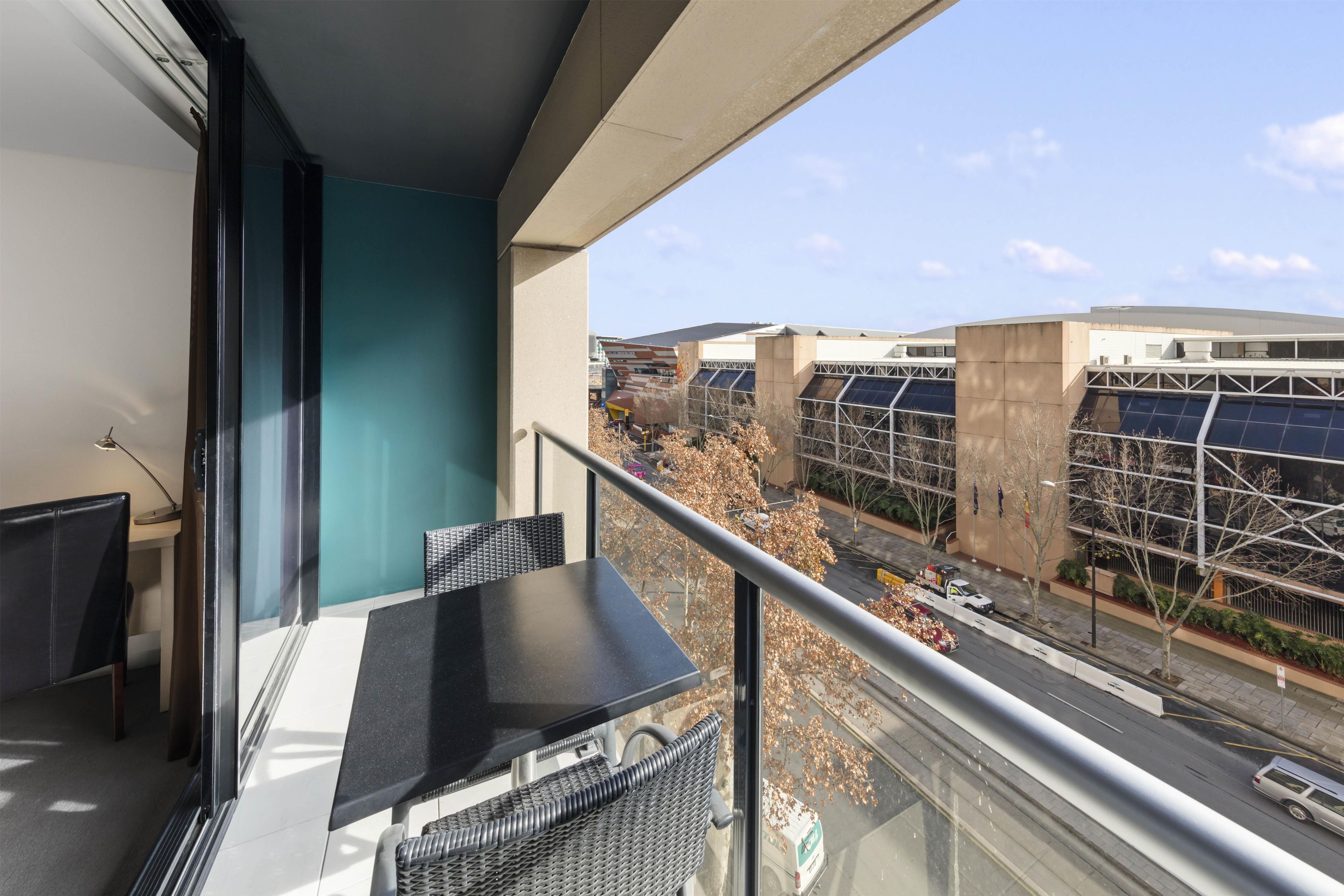 Un balcon avec une table et des chaises, donnant sur une rue bordée d’immeubles et offrant une vue sur un ciel dégagé.
