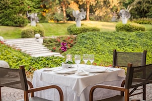 Un espacio al aire libre para comer con una mesa dispuesta para dos personas, rodeado de follaje y estatuas.