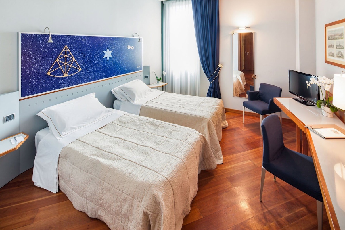 Quarto de hotel com duas camas, uma escrivaninha com cadeira, televisão e uma janela grande com cortinas.