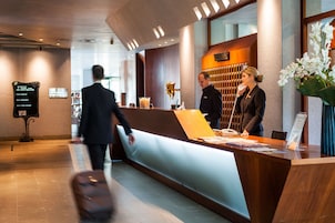 Zona de recepción de un hotel con un mostrador de madera, un separador de vidrio y un cartel que enumera los tipos de habitaciones.