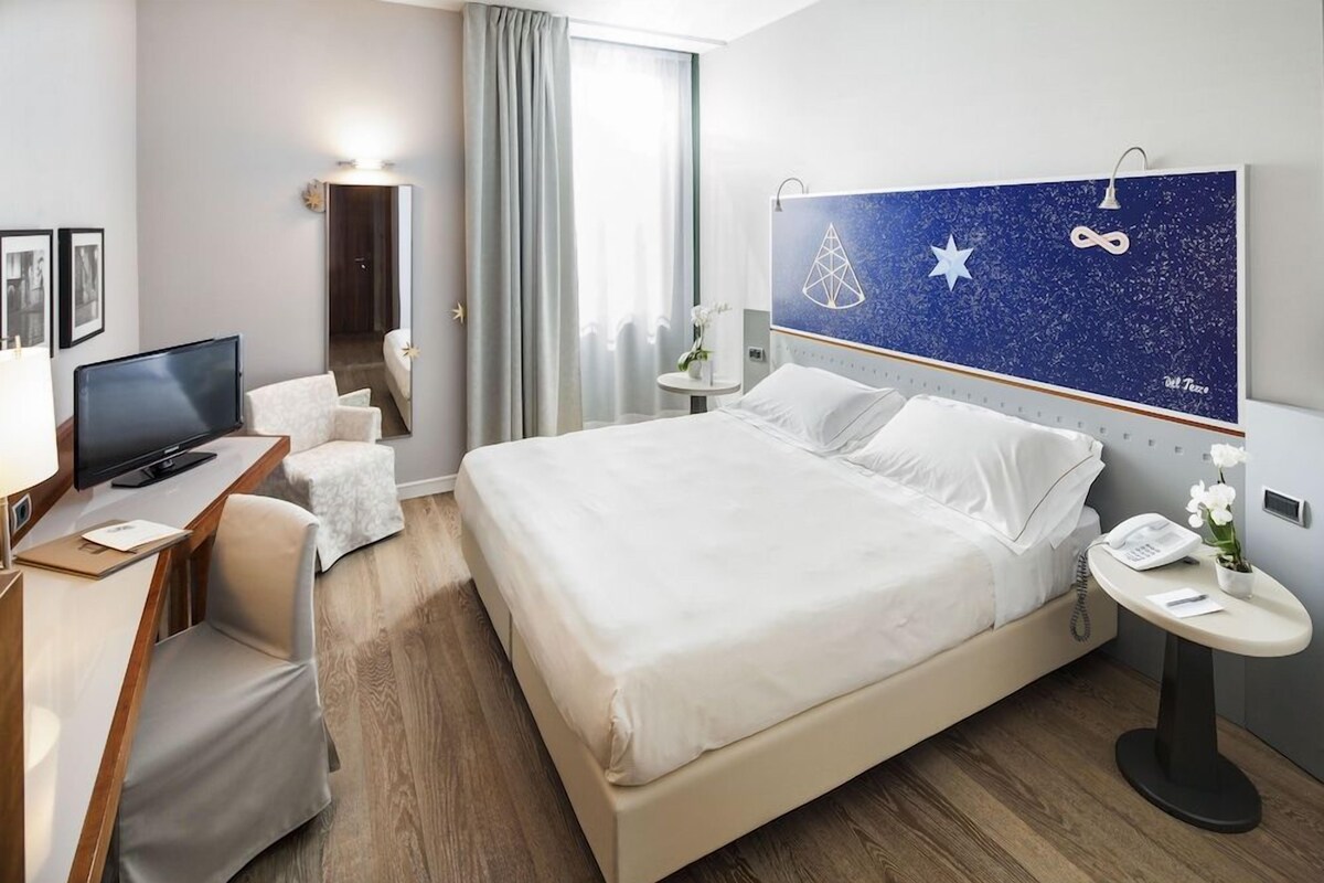 Hotellihuone, jossa on sänky, työpöytä, jossa on televisio, pieni pöytä, jossa on puhelin, ja peili.