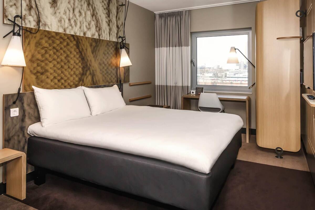 Una habitación de hotel moderna con una cama grande, un escritorio, una silla y vistas a la ciudad por la ventana.