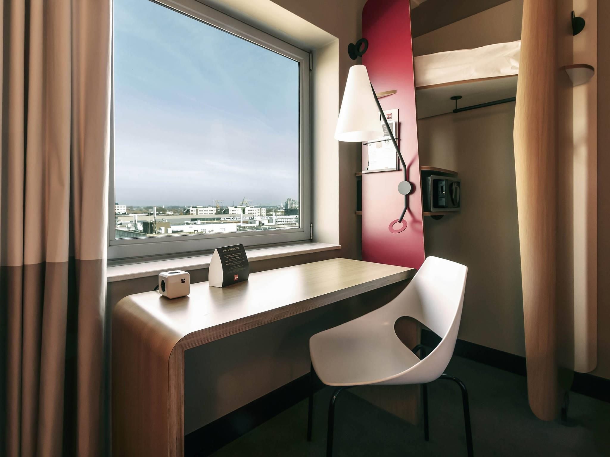 A hotel room with a desk, chair, lamp, and a view of the city through a large window.