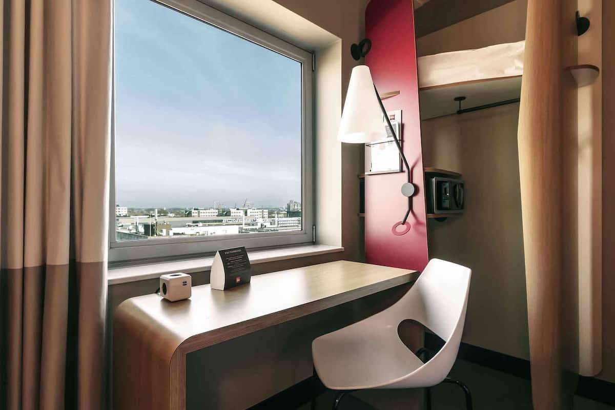 Une chambre d’hôtel équipée d’un bureau, d’une chaise, d’une lampe et offrant une vue sur le paysage urbain par la fenêtre.