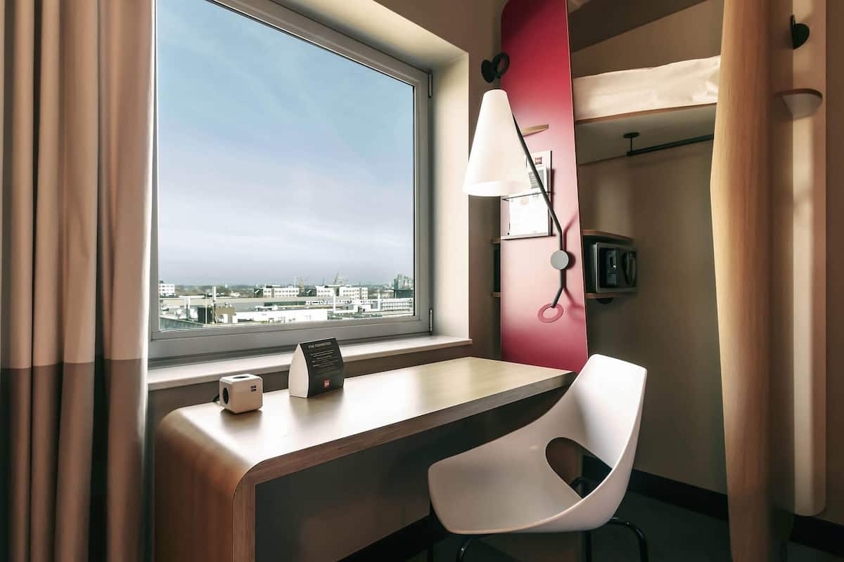 Habitación de hotel con escritorio, silla, lámpara y vistas a la ciudad a través de un ventanal grande.