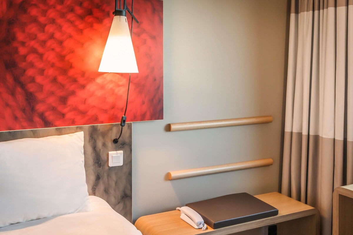 Habitación de hotel con una cama, una mesita de noche con un teléfono y un libro, una lámpara de pared y una pared texturizada de color rojo.