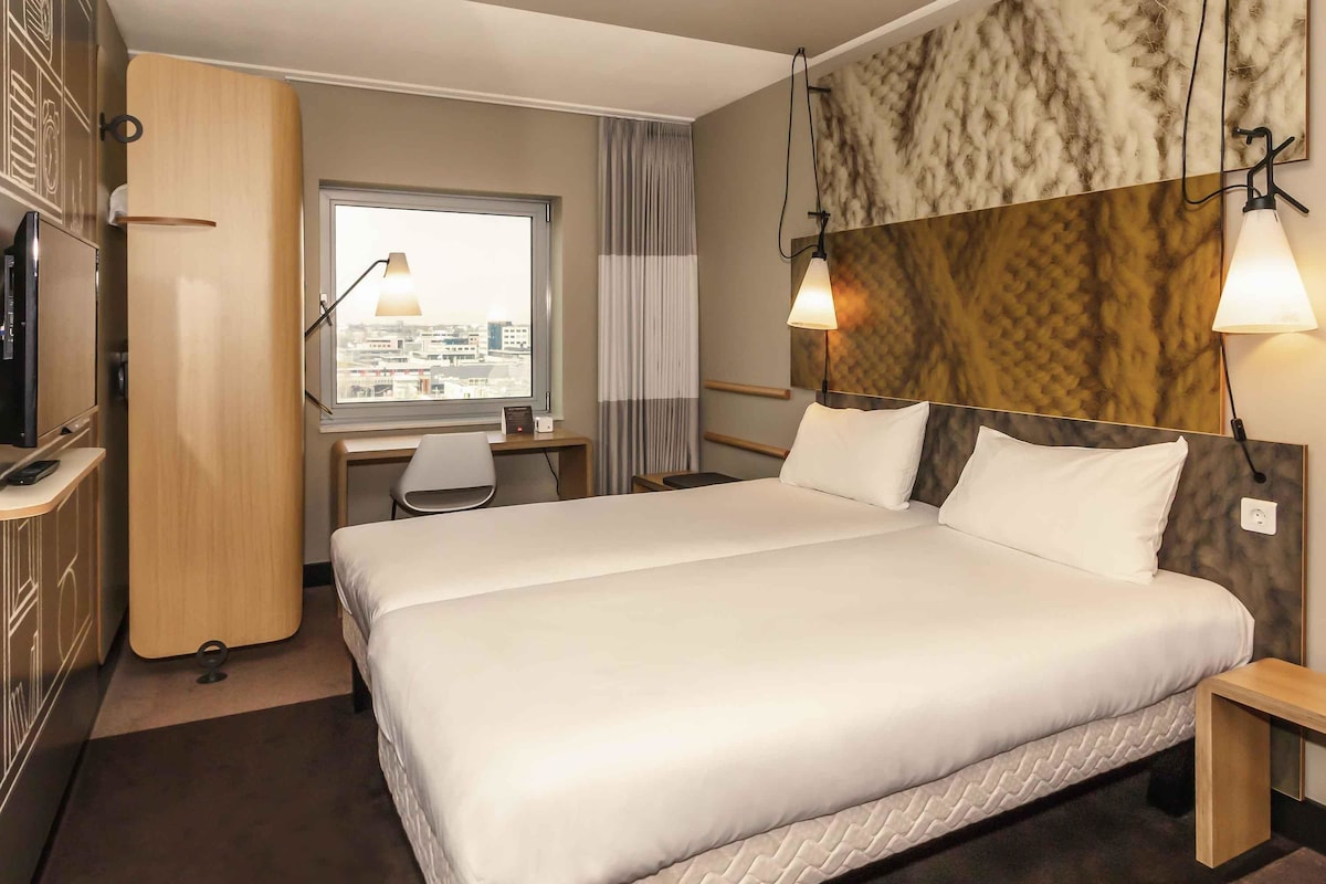 A hotel room with a large bed, a desk, a chair, a wardrobe, and a window with a city view.