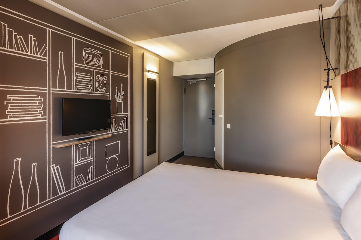 Een hotelkamer met een groot bed, een flatscreen televisie aan de muur en een modern design met geometrische wanddecoratie.