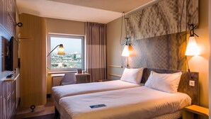 Habitación de hotel con dos camas, un escritorio, una silla, un ventanal con vistas a la ciudad y una pared decorada con mármol.