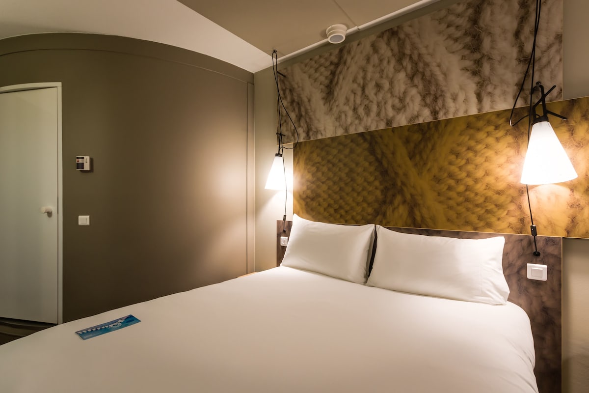 A hotel room with a bed, a headboard with a textured pattern, and two wall-mounted lamps.