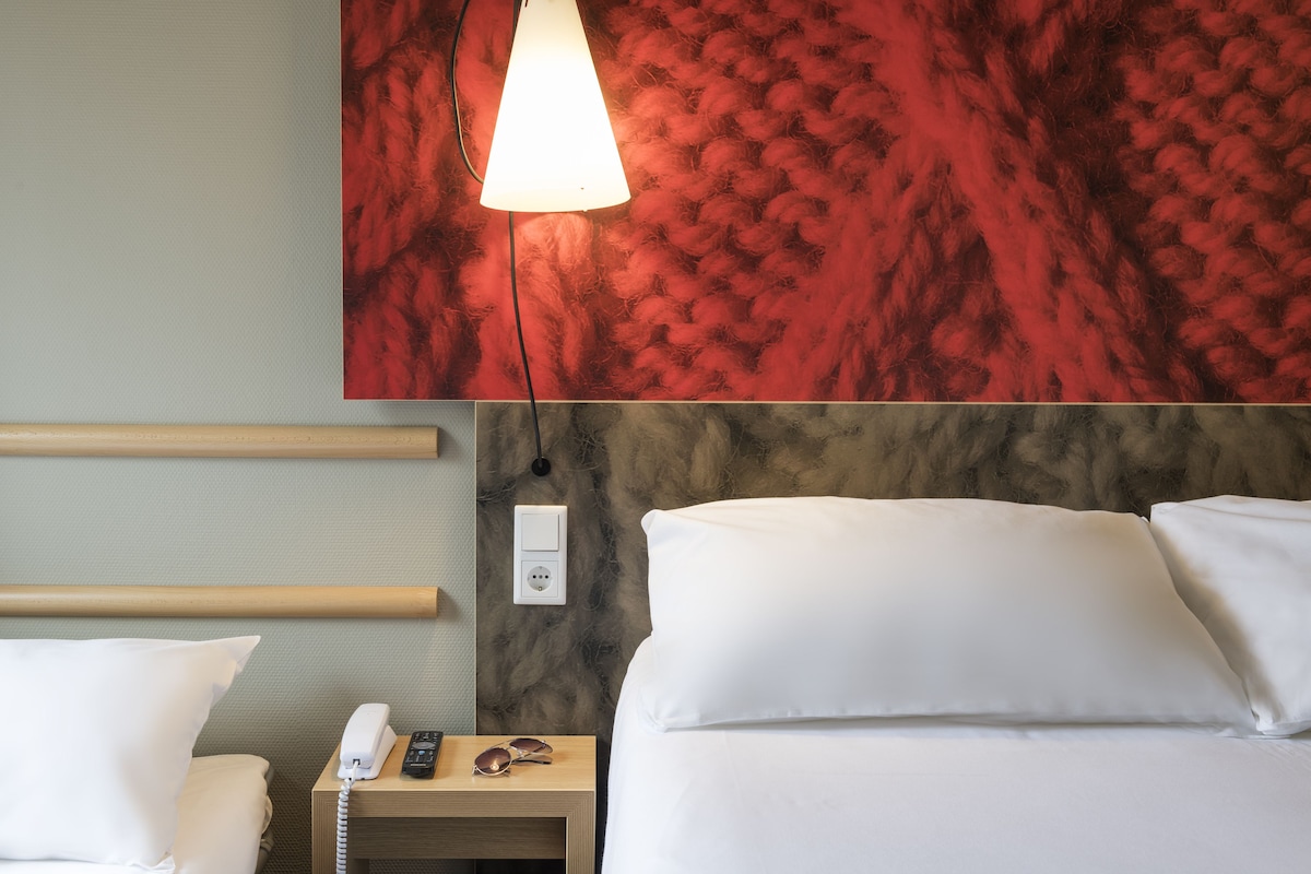 A hotel room with a bed, bedside table, lamp, and a wall with a textured red pattern.