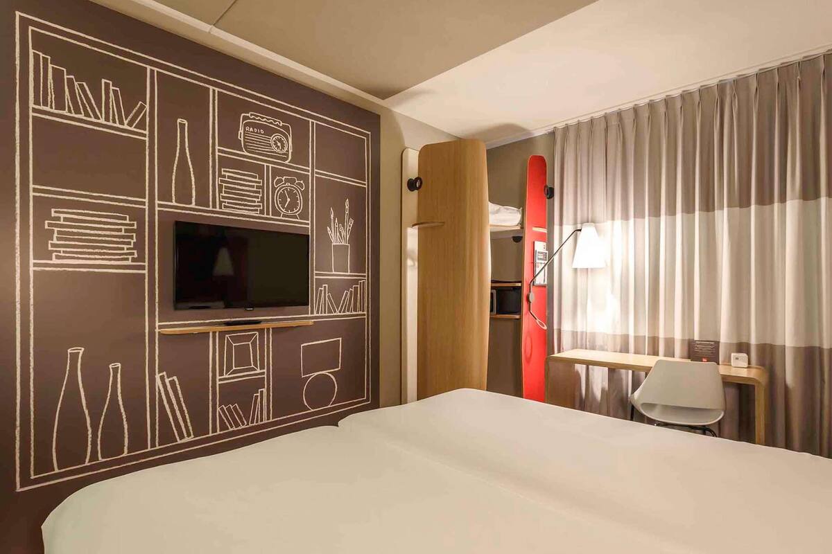 Habitación de hotel con cama, televisión montada en la pared, escritorio con silla y ventana con cortinas.