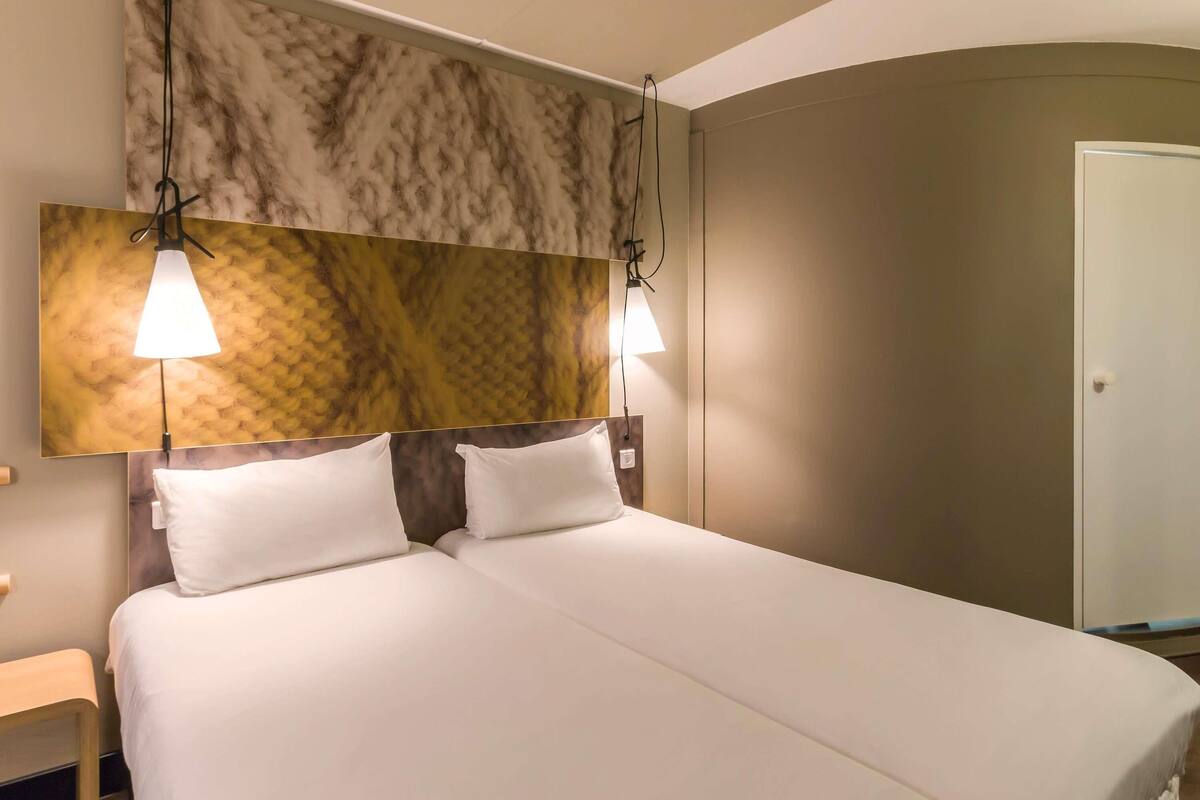Una habitación de hotel con cama, dos lámparas de noche, cabecera con diseño texturizado y una puerta.