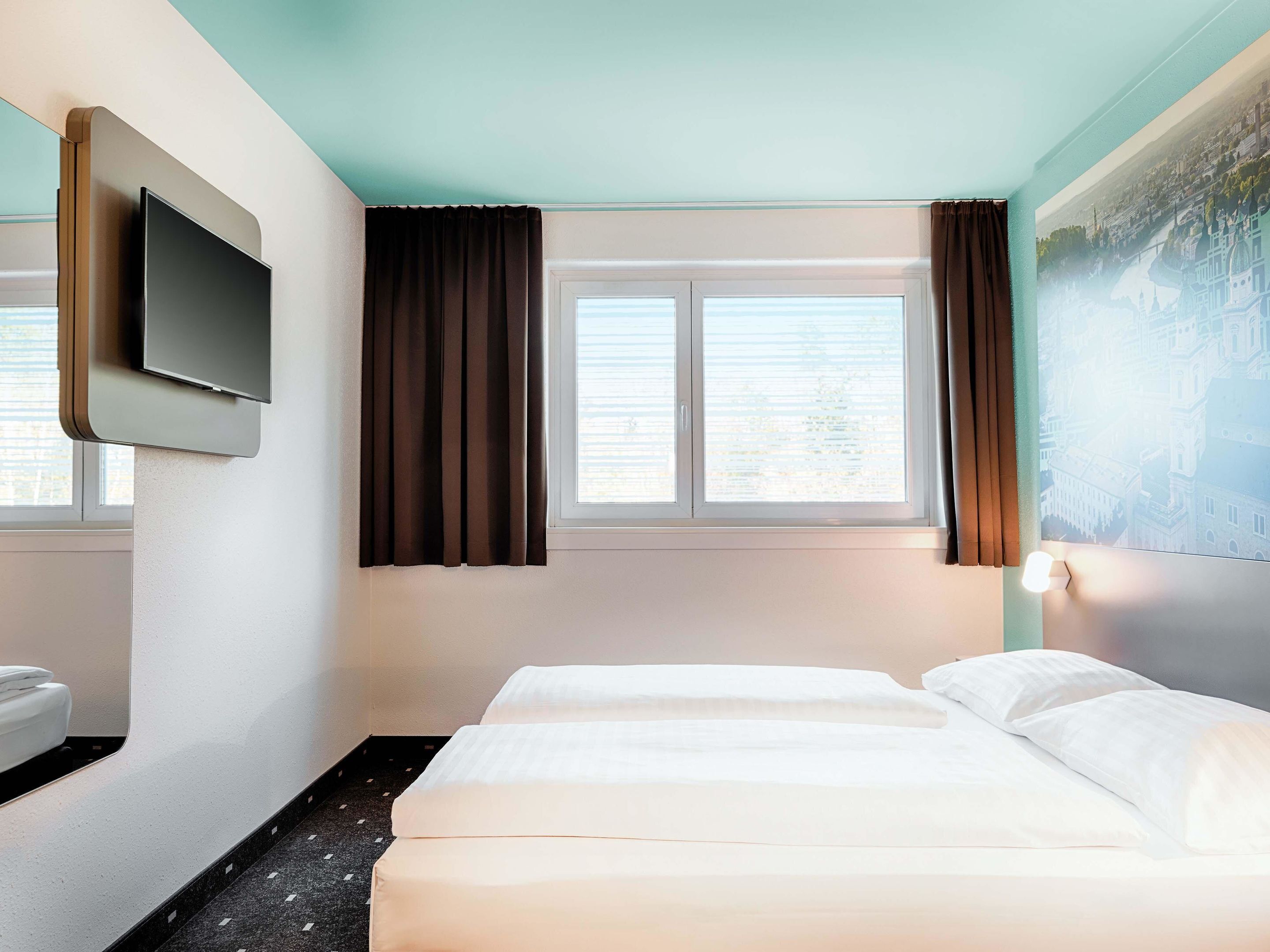 A hotel room with two beds, a flat-screen TV mounted on the wall, a window with curtains, and a painting on the wall.