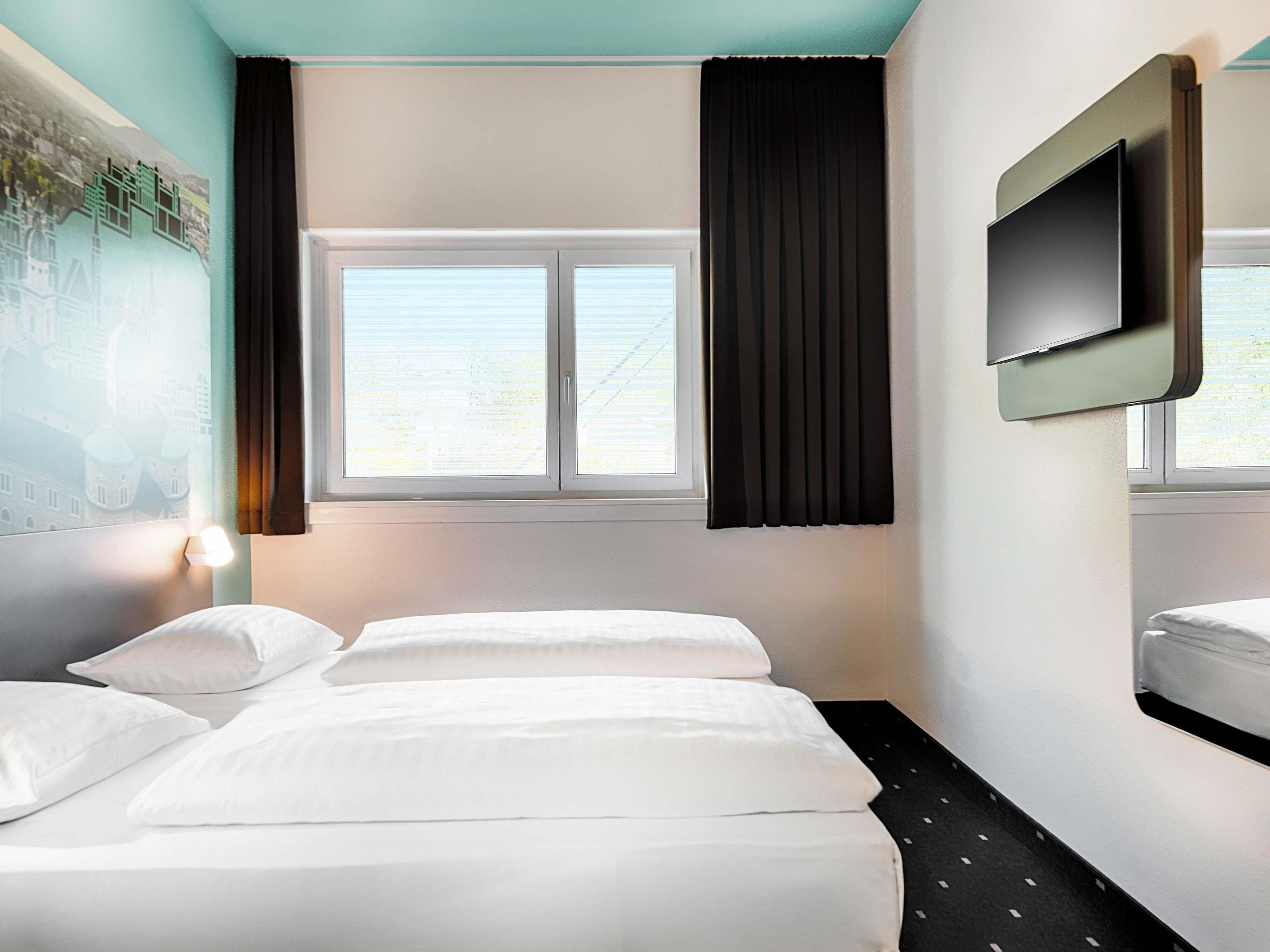 Une chambre d’hôtel avec deux lits, une grande fenêtre avec des rideaux noirs, un téléviseur fixé au mur et une lampe fixée au mur.