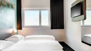 Une chambre d’hôtel avec deux lits, une grande fenêtre avec des rideaux noirs, un téléviseur fixé au mur et une lampe fixée au mur.